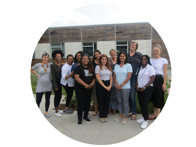 Photograph of fourteen diverse women smiling, posing outdoors in front of a building.