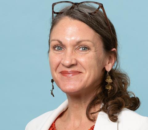 Kat Theall, smiling, with glasses on her head, wearing a white blazer against a blue background.