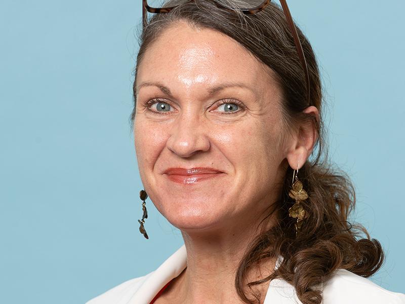 Kat Theall, smiling, with glasses on her head, wearing a white blazer against a blue background.